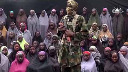A man in military camouflage with his face covered and holding an AK-47 assault rifle stands in front of a 50 or so scared-looking girls in hijabs that cover their bodies