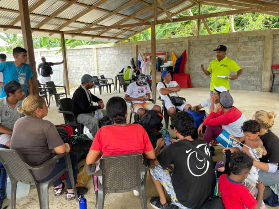 A group of people of different ages with children sit listening to a man in an open-sided shelter