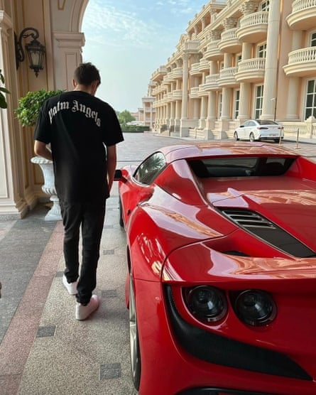 A man wearing a Palm Angels shirt stands next to a red luxury car