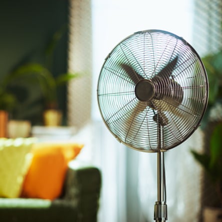 A modern stand fan with spinning blades is prominently featured, providing refreshing air circulation in a comfortable living room with a green velvet sofa and natural light.