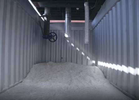 The interior of a metal shipping container with lots of white pebbles piled up at one end and two silver pipes and one dark metal pipe with a valve wheel.