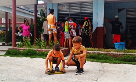 Two children play in a yard while a group of adults chat in the shade of a building