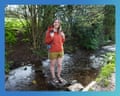 A woman in an orange jacket and yellow shorts stands on a rock in a shallow stream, wearing sandals and carrying a backpack.