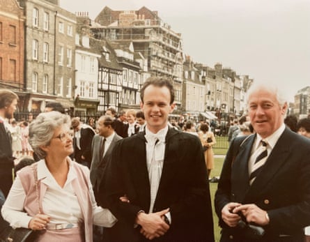 Chris Moore wearing a black gown and white shirt and white bow tie, standing with his mother on his right, her arm linked with his, and his father to his left, standing on a lawn outside university buildings