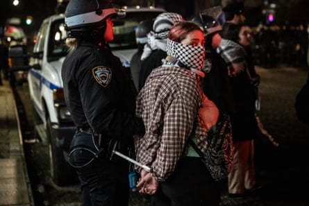 Night shot of woman whose face is wrapped in keffiyeh with hands ziptied behind her back behind led by police.