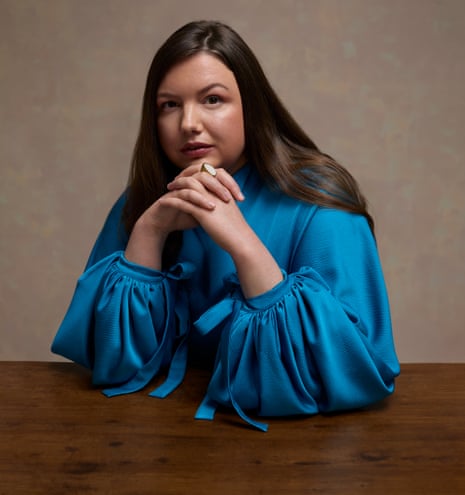 Hannah Murray wears a vibrant blue dress with voluminous sleeves and sits at a wooden table with her hands clasped