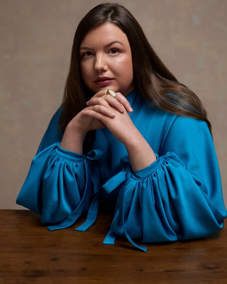Hannah Murray wears a vibrant blue dress with voluminous sleeves and sits at a wooden table with her hands clasped