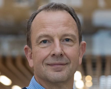 Portrait of Jason Tarry. He is in his late 50s with receding light brown hair and wears a blue and white checked shirt under a dark jumper.