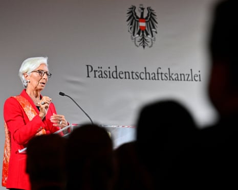 Woman with grey hair and spectacles, dressed in red blazer and orange scarf addresses audience in Vienna.