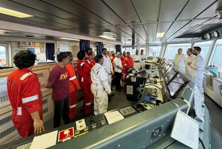 People wearing red and white boiler suits gathered around the control panels on the bridge of a ship.