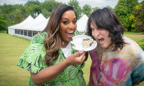 Alison Hammond, with long hair and in a short-sleeved dress, next to Noel Fielding in a field with tents behind them, both pretending to bite a plate with a slice of cake