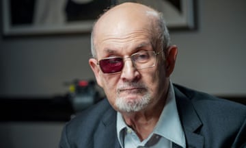 Salman Rushdie in a shirt and jacket, with a moustache and goatee and glasses that have the righthand frame darkened, looking serious