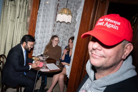 man in red ‘make veterans great again’ baseball cap smiles as people sit at table behind him
