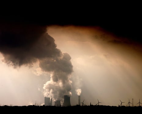 Ominous smoke and vapour rise from the cooling towers and chimneys of a power plant, with smaller wind turbines in the foreground