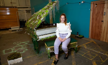 Kirsten Little, with long hair and wearing glasses and a shirt with flouncy-shouldered sleeves, smiles as she sits at a baby grand piano decorated with living moss in the Make CIC workshpace