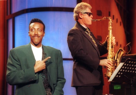 a man in su glasses plays the saxophone as another man points at him and smiles