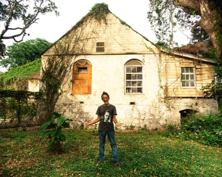 Williams stands in the botanic gardens in front of a whitewashed stone building; he has his arms outstretched. He is thin and has a grey beard; he wears a dark grey T-shirt with an image of Malcolm X on the front, a black woollen hat and blue jeans