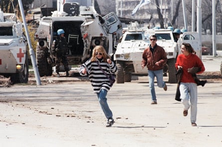 A young woman in a striped jumper, jeans and sunglasses, followed by another in a red jumper and with a man in a brown anorak behind them, run across a concrete strip; white UN armoured vehicles and soldiers stand behind them.