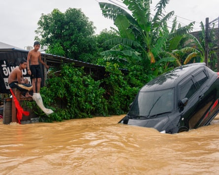 Two boys look at a black Mini car submerged in passing floodwater