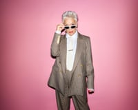 Denise Welch pictured from above the knee, wearing a pale-brown trouser suit, a white shirt and tie with a tie-pin, and black, white-rimmed sunglasses, which she is holding with her right hand on the bridge of her nose while looking at the camera, against a rose-pink background