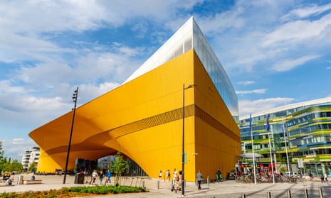 A low angle photo shows a bright yellow building looming towards the sky, its sharp corner and edges contrasting with a front side that curves down to a recessed entrance