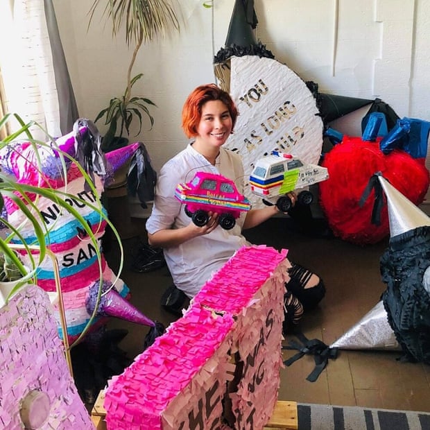 A woman with short red hair is surrounded by colorful piñatas as she holds two smaller ones in her hands.