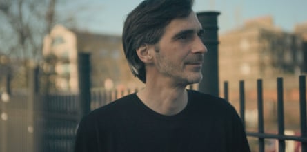 A man with neat side parted hair and wearing a black jumper smiles as he stands nect to some metal railings
