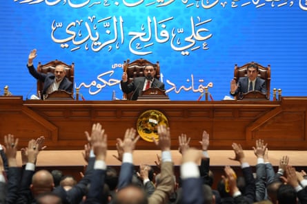 A parliamentary session owhere Al-Zaidi and most members of his cabinet received a vote of confidence. People lift their hands in the room front of a panel of three men in suits