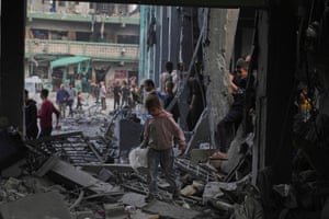 A child in the foreground walks over debris; other people stand in a courtyard in the background