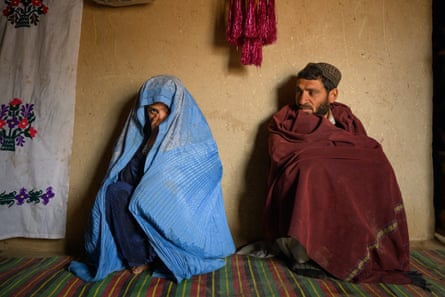 A man wrapped in a cloak sits against a wall next to a girl wrapped in a chador with just her face showing