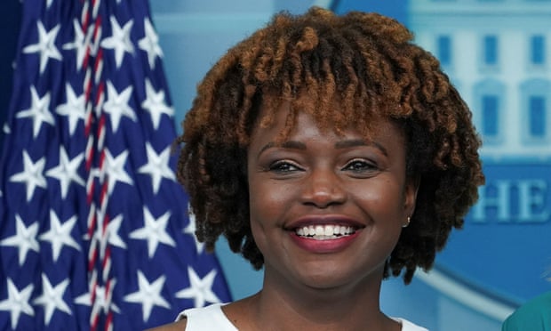 Karine Jean-Pierre stands in front of an American flag with the presidential seal in the background.