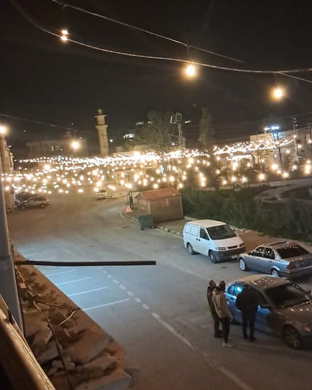 A road next to a lit-up area, at night, with small fairy lights crisscrossing the area