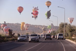 Brightly coloured hot air balloons, some in the shape of cartoon figures and creatures, float in the sky above cars on a busy road