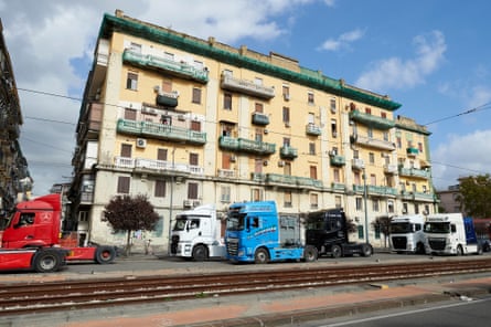 A line of red, white and blue truck cabs stands stationary on a street in Naples in front of a slightly dilapidated yellow stone apartment building.