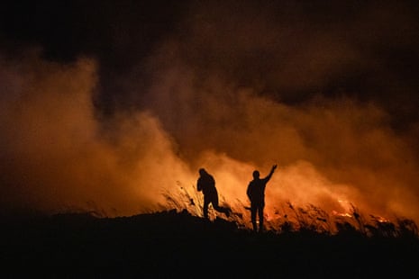 Two figures are silhouetted by wind-driven flames and smoke