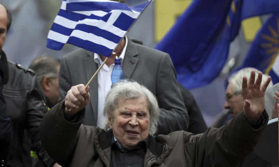 Theodorakis in old age, holding both hands up to salute the crowd, with a blue and white Greek flag on a stick in one hand