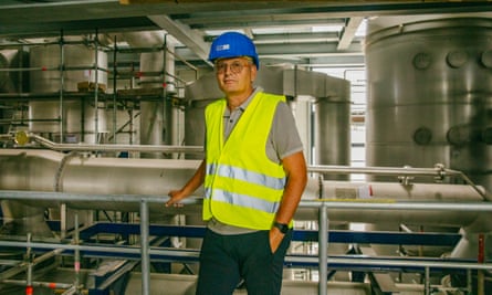 A man in a blue hard hat and yellow hi-vis vest poses for a photo with one hand on a safety rail in front of factory equipment