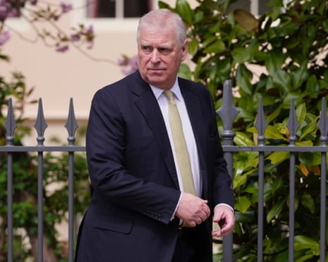 Andrew Mountbatten-Windsor walks near Windsor Castle in front of black railings and leafy greenery. He wears a dark jacket, white shirt and gold tie.