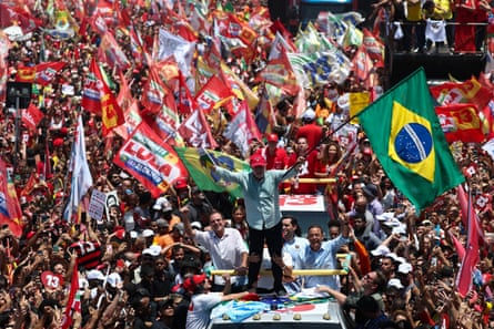 Man in turquoise button-down and red cap stands on top of vehicle while holding Brazilian flag in front of large crowd of people in streets