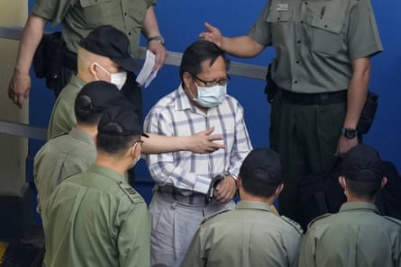 Albert Ho is escorted by a group of correctional services officers wearing khaki shirts, who surround him; he is a small man of about 70 and is wearing a white shirt with large grey checks and grey trousers plus a blue face mask.