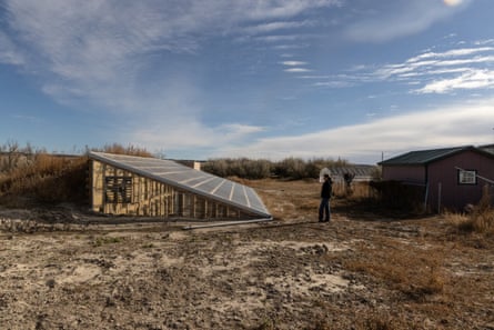 A women stands beside a wooden greenhouse sunk into the ground with a slanted roof