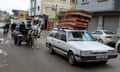 A car drives with several mattresses strapped to the roof, followed by two people on a donkey-drawn cart and others on bicycle or foot