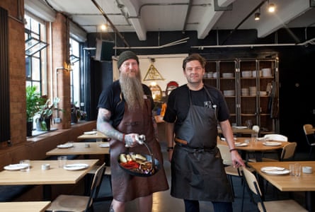 Two men in black T-shirts and aprons in a kitchen. One has a long grey fuzzy beard. The other has short dark hair