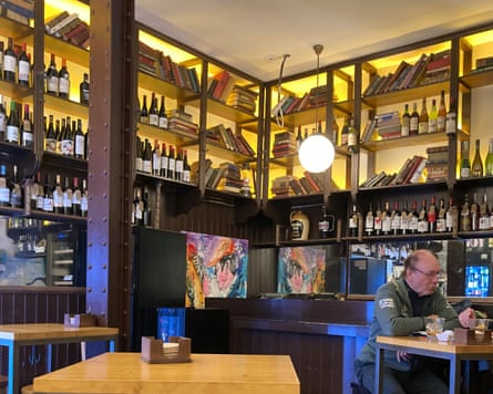 A man sitting on a stool at a light wood table with illuminated shelves of wine and books behind him