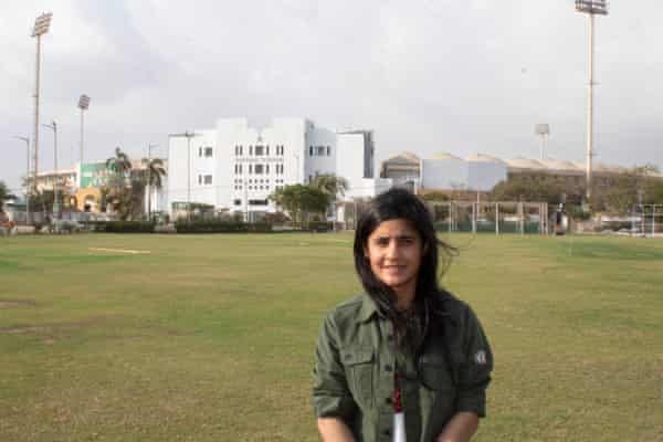A woman stands on a cricket pitch with a large white pavilion in the background
