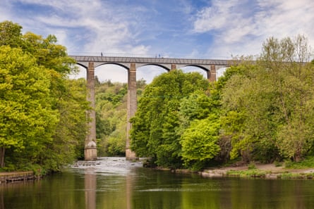 Tiny figures walk across a high, arched aqueduct over a river and surrounded by trees