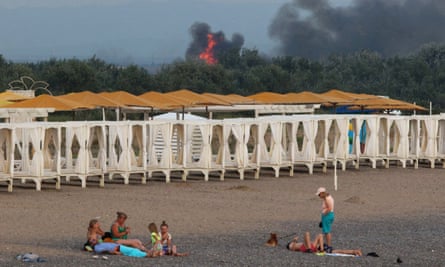 People on a beach as smoke and flames rise in the distance after explosions at a Russian military airbase in Novofedorivka, Crimea
