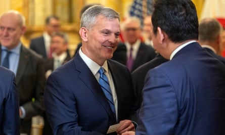 A white man with gray hair wearing a blue suit shakes another man’s hand and smiles.