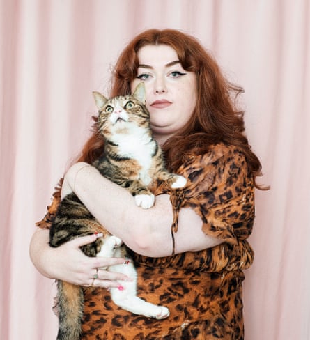 Ione Gamble wearing an animal print dress, holding her tabby cat, a pale pink curtain behind