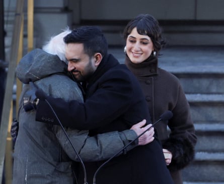 Rama Duwaji, in brown funnel neck, watches Bernie Sanders embrace her husband, Zohran Mamdani, at his inauguration ceremony